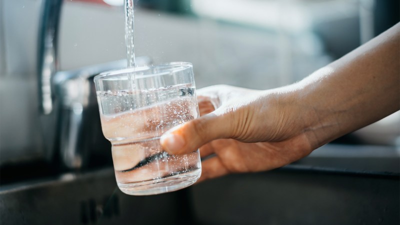 A stock image of a person holding a glass under a running faucet. The cup is filling with drinking water.